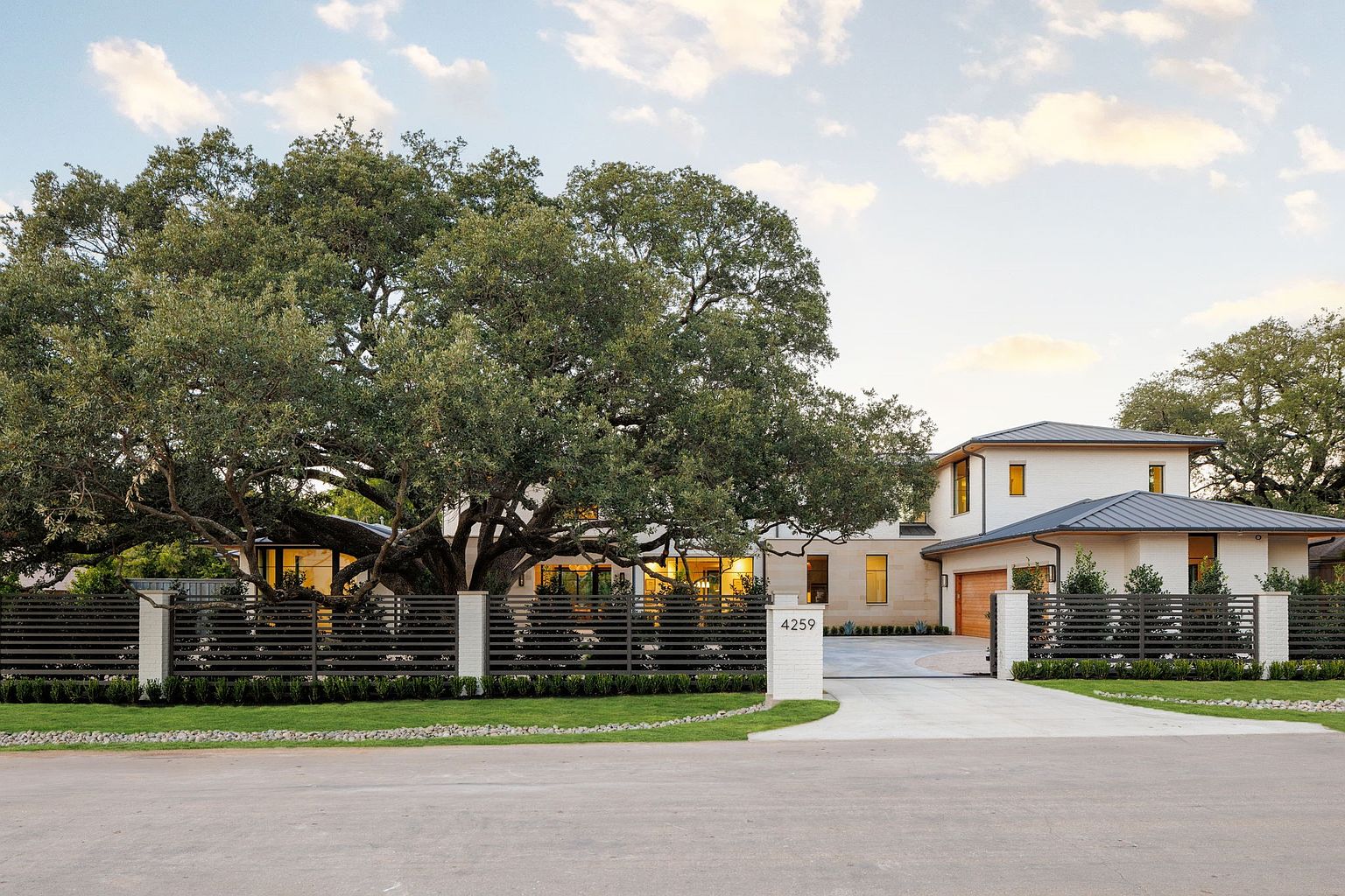 This is a front exterior view of a modern home featuring a large tree in the front yard. The house has a light-colored brick facade, a dark metal roof, and a horizontal slat fence with brick pillars. The driveway leads to a garage, and the address "4259" is displayed on one of the pillars.