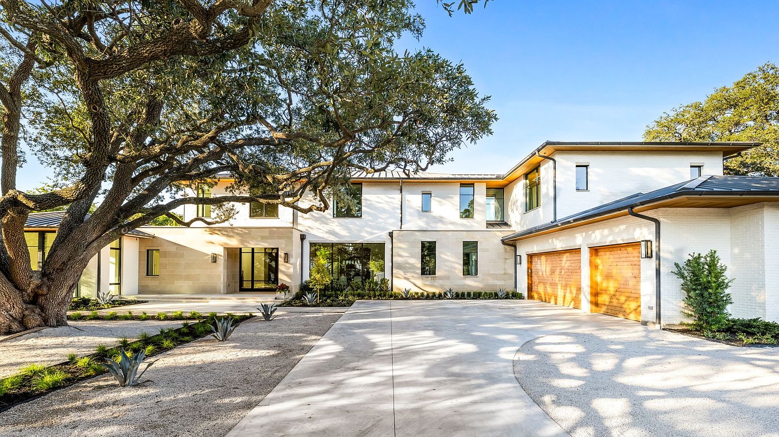 This is a front exterior view of a modern, two-story home with a combination of stone and white brick facade. The property features a well-manicured front yard with gravel pathways and mature trees providing shade. A concrete driveway leads to a two-car garage with wooden doors, enhancing the home's curb appeal.