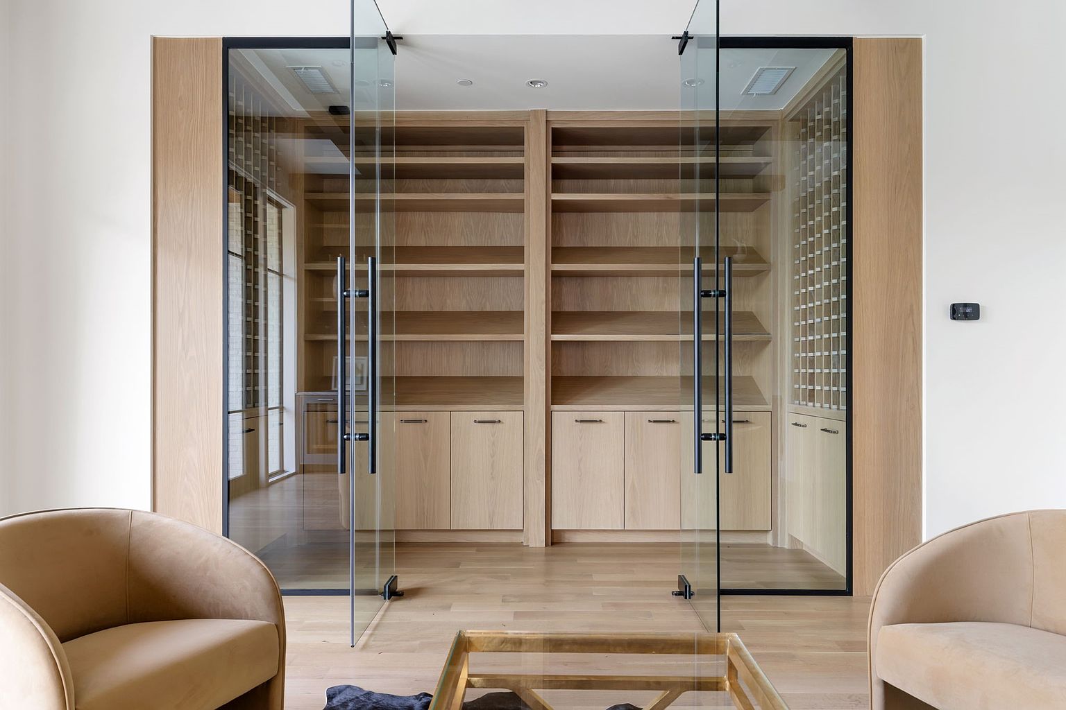This interior shot showcases a sophisticated office or study space, featuring custom built-in shelving and cabinetry in a light wood finish. Glass doors with black frames provide a modern touch, while two armchairs and a glass coffee table create a comfortable seating area. The room exudes a sense of refined elegance and functionality, perfect for a professional or personal workspace.