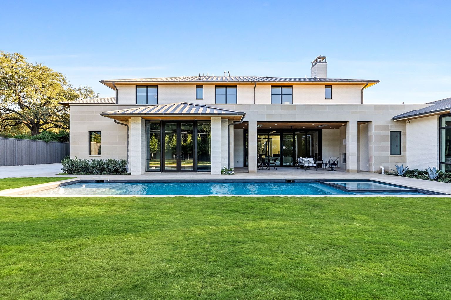 This is a rear exterior view of a luxurious modern home, showcasing a pristine pool and spa area. The house features a light-colored brick facade, large glass doors, and a covered patio with outdoor seating. The well-manicured lawn extends to the pool, creating an inviting and serene outdoor living space.