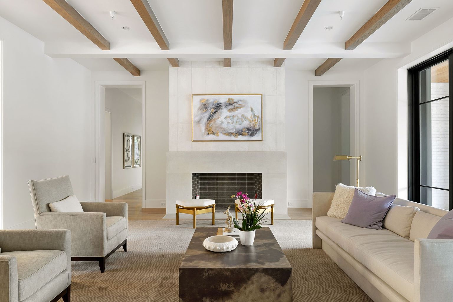 This is an interior shot of a living room featuring a fireplace with a light stone surround and a modern abstract painting above it. The room is decorated in neutral tones with wooden ceiling beams, a light-colored sofa, and two armchairs. The space is well-lit and appears spacious, creating a comfortable and inviting atmosphere.