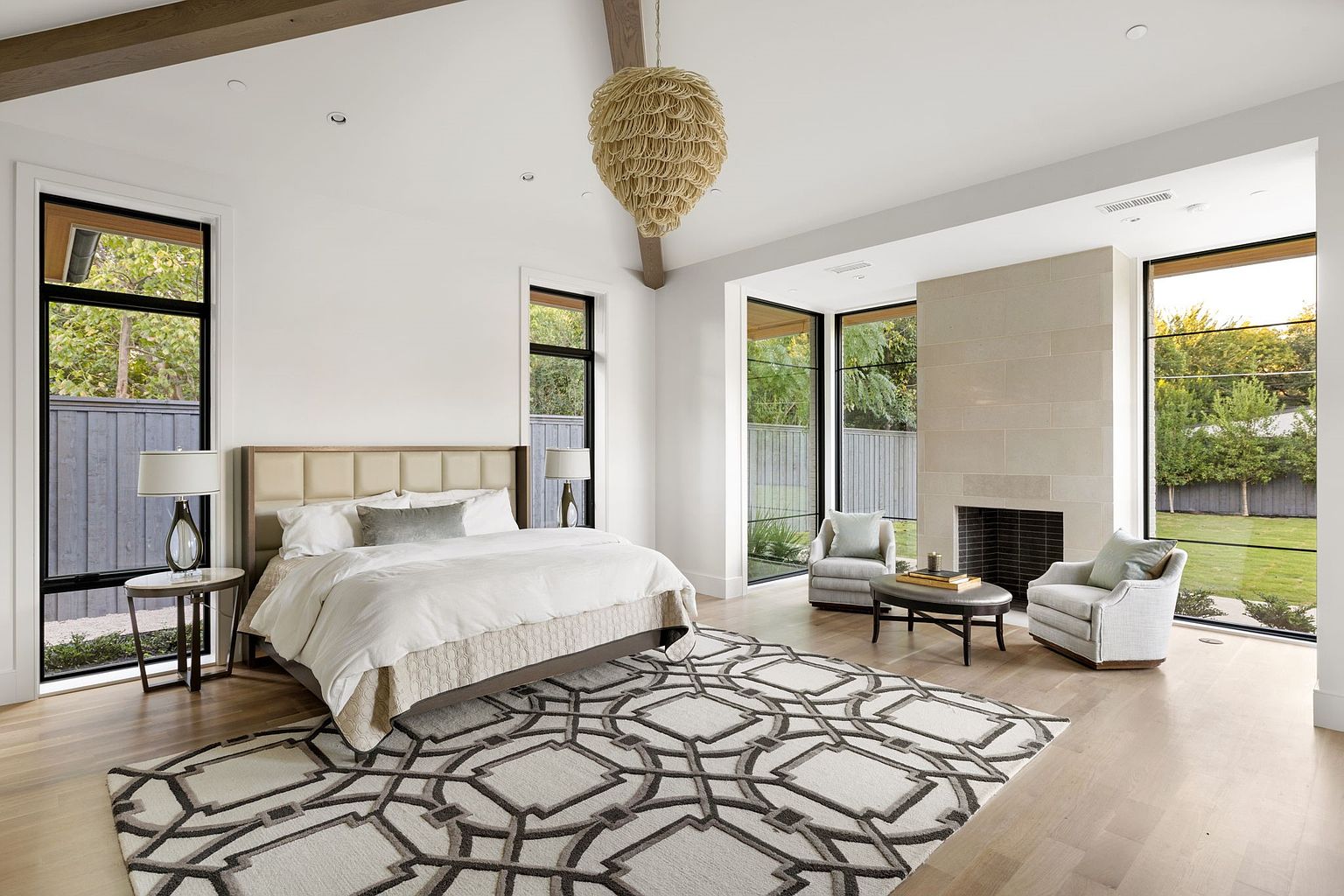 This is a bright and spacious primary bedroom featuring a large bed with a neutral-toned headboard, a geometric patterned rug, and a modern fireplace. Large windows provide ample natural light and views of the outdoors, while a unique chandelier adds a touch of elegance. The room is styled with a minimalist approach, creating a serene and inviting atmosphere.