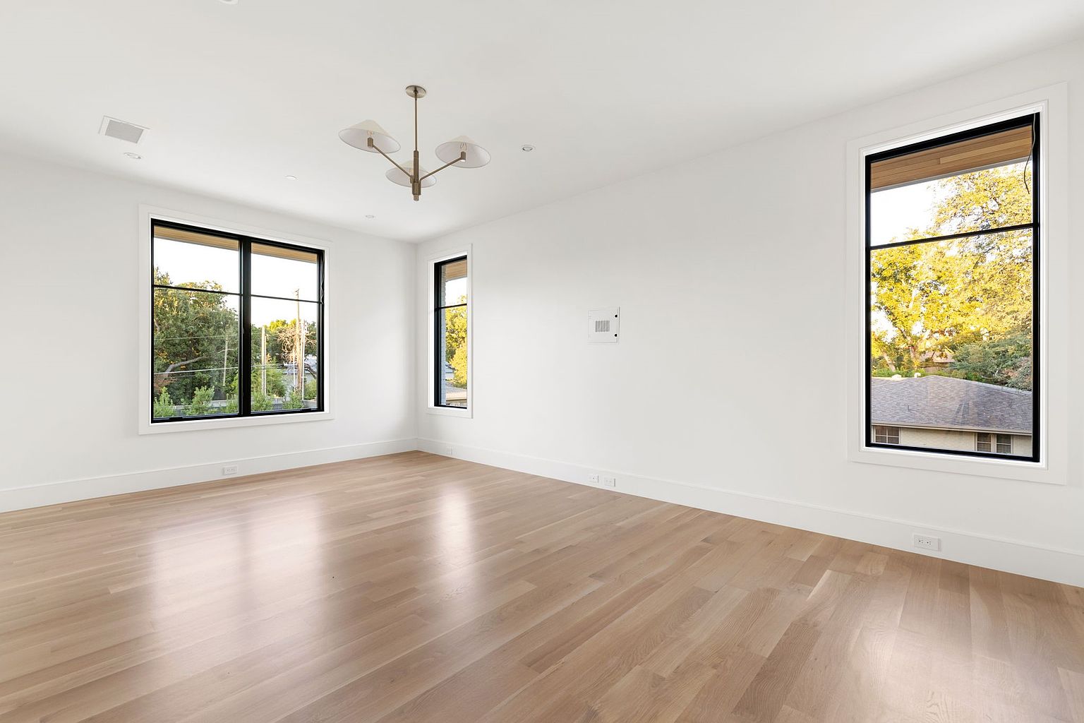 This is a bright and airy living room featuring hardwood floors, white walls, and large black-framed windows that provide ample natural light. A modern chandelier hangs from the ceiling, adding a touch of elegance. The room is spacious and uncluttered, creating a clean and inviting atmosphere.