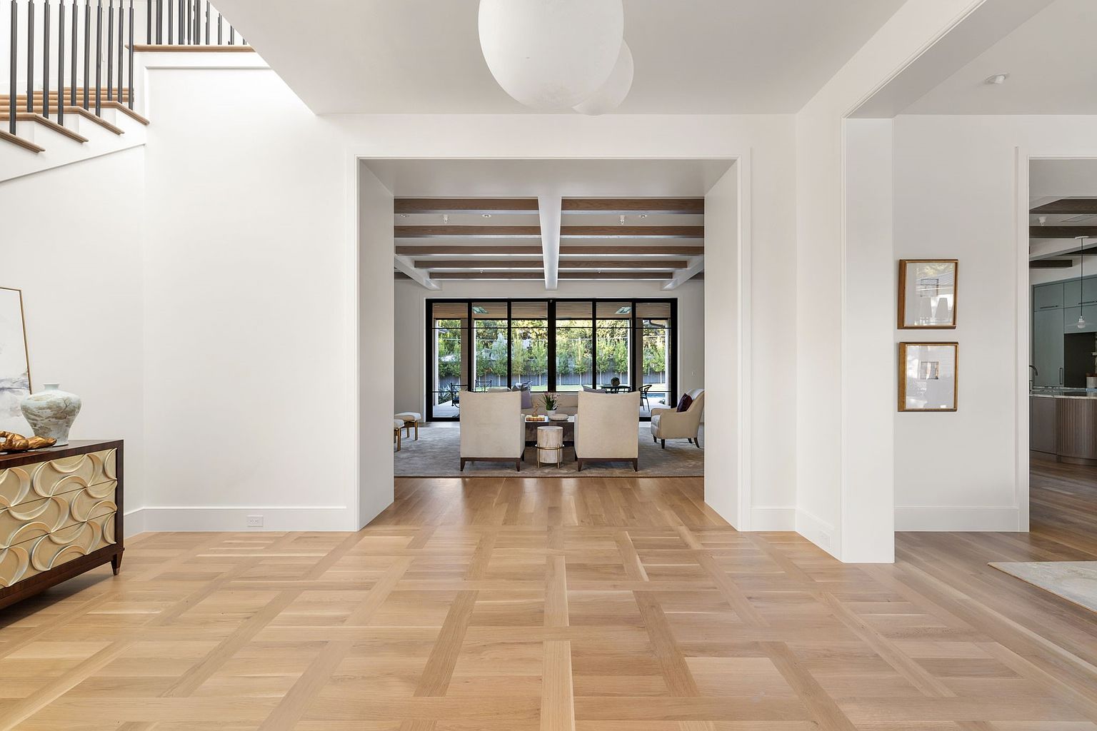 This interior shot showcases a grand hallway with light wood flooring in a geometric pattern, leading to a bright living area visible through a large opening. The hallway features white walls, a staircase on the left, and framed artwork on the right, creating a clean and elegant transition space within the home. The perspective is from the entrance, looking into the house.