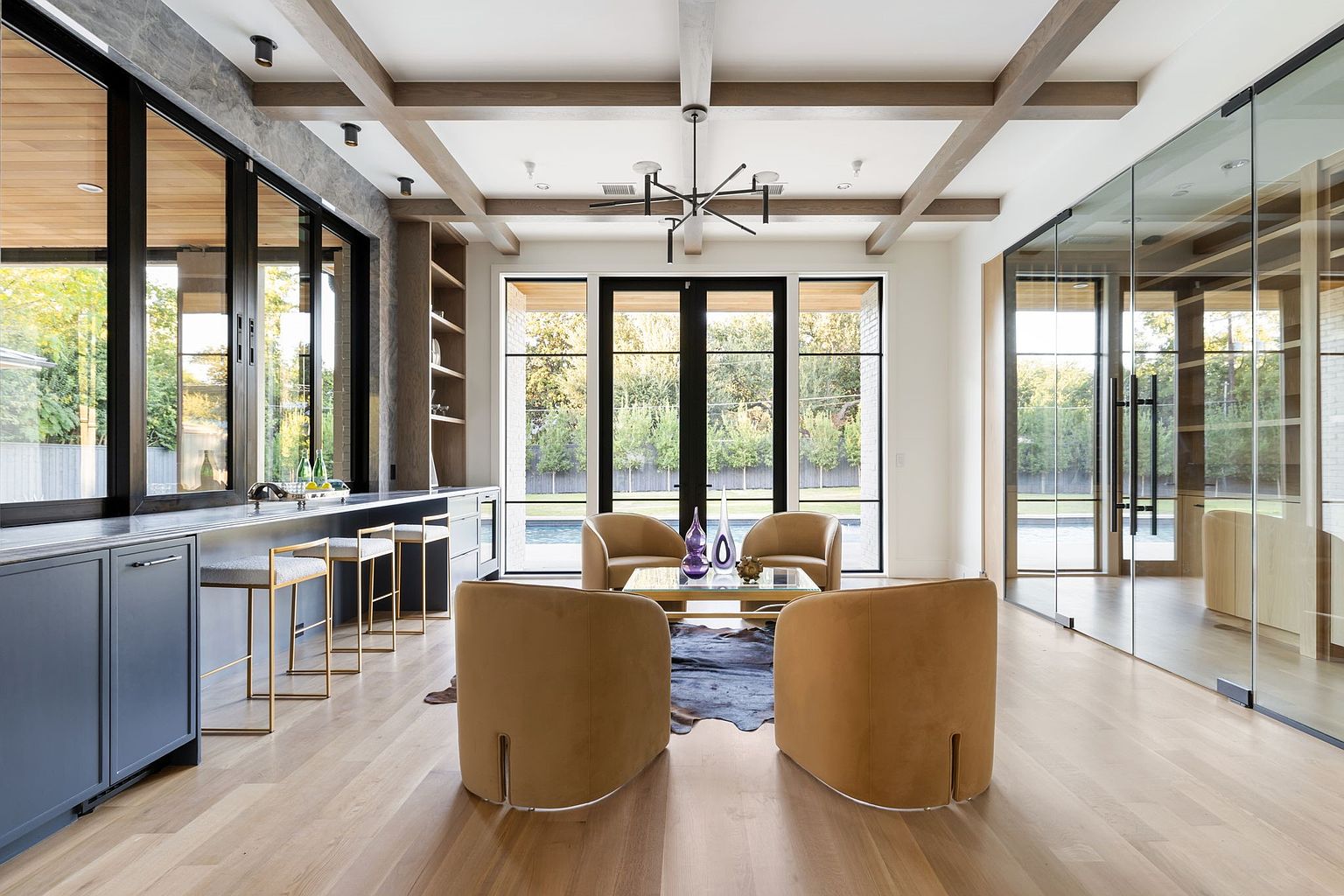 This interior shot showcases a modern living room with a focus on natural light and contemporary design. The room features hardwood floors, a coffered ceiling with wooden beams, and large windows offering views of the outdoors. Four tan-colored armchairs are arranged around a glass coffee table on a cowhide rug, creating a cozy and inviting seating area.