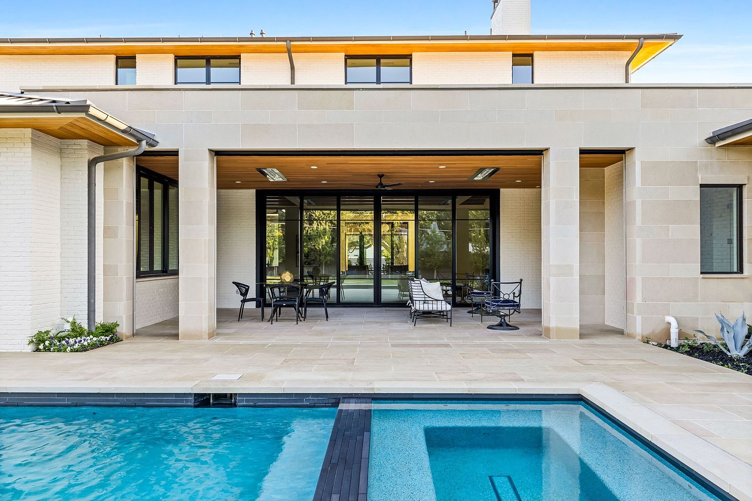 This image showcases a luxurious outdoor space featuring a modern pool and spa area. The pool has a sleek design with a dark tile accent, and the adjacent patio is furnished with stylish outdoor seating. The architecture of the home is contemporary, with clean lines and a neutral color palette, creating an inviting and sophisticated atmosphere.