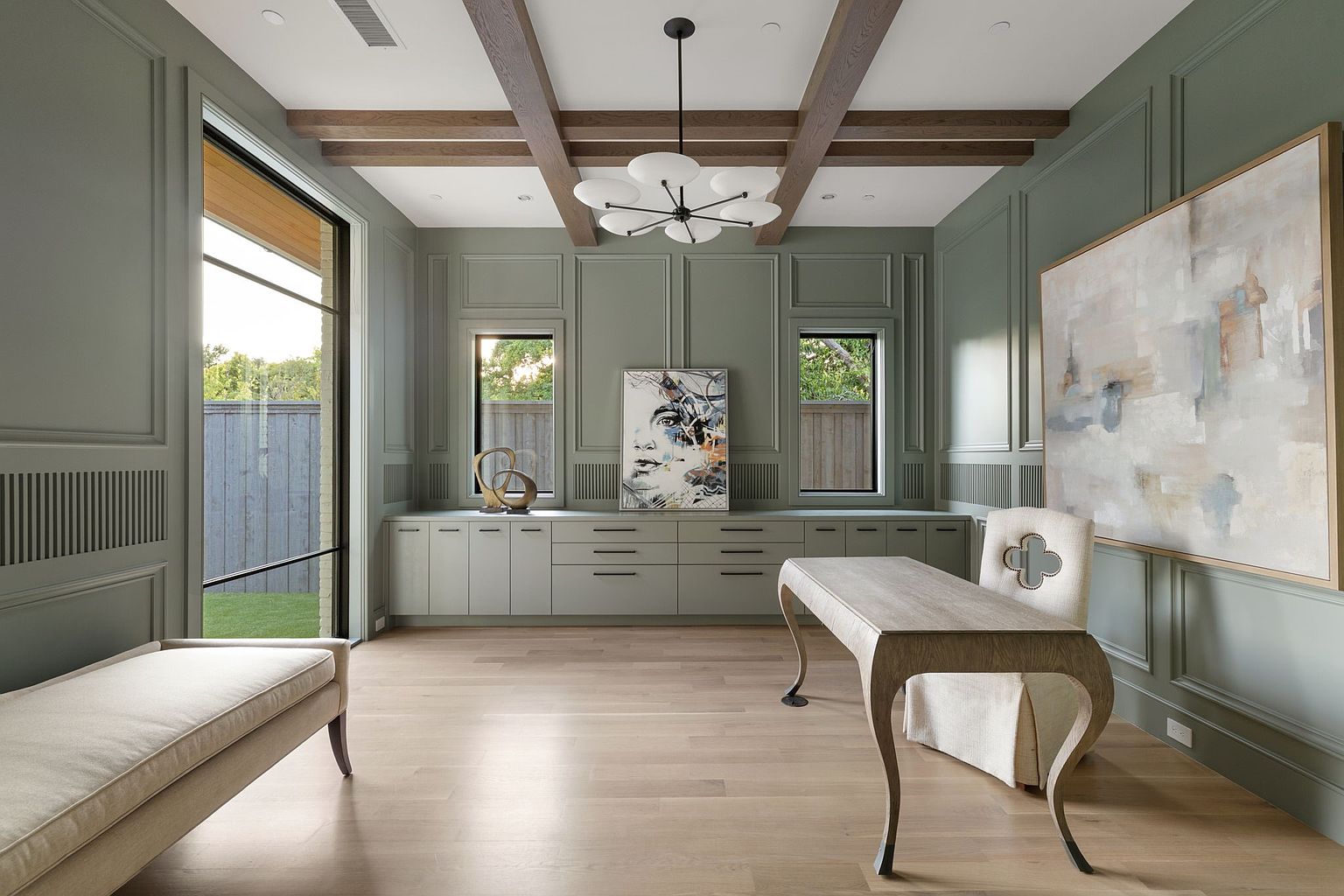 This is an interior shot of a sophisticated home office featuring sage green walls with decorative molding, light wood flooring, and exposed wooden beams on the ceiling. The room is furnished with a light-colored desk, a chair with a unique clover design, and a chaise lounge, complemented by modern artwork and a stylish chandelier. Large windows provide natural light and a view to the outside, creating a serene and elegant workspace.