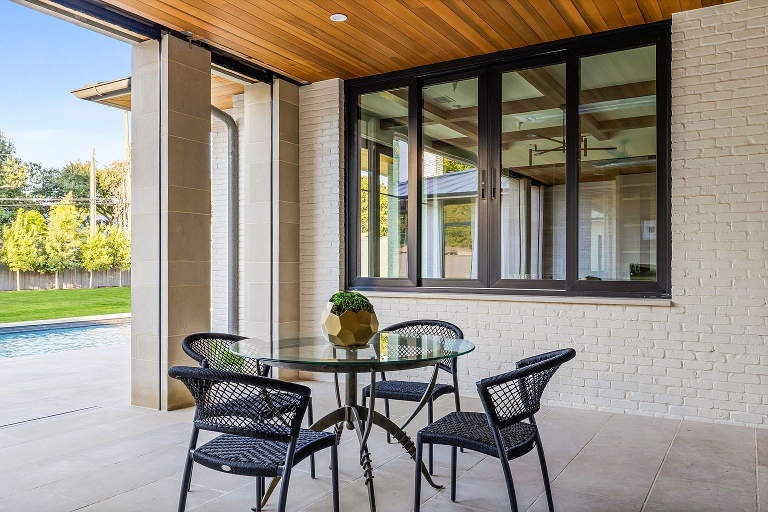 This image showcases a stylish outdoor patio area, featuring a glass-topped table surrounded by four black woven chairs. The patio is adjacent to a modern home with large windows and a brick exterior, and the space is covered by a wooden ceiling. The scene offers a glimpse of a well-maintained lawn and pool, suggesting a luxurious and relaxing outdoor living space.