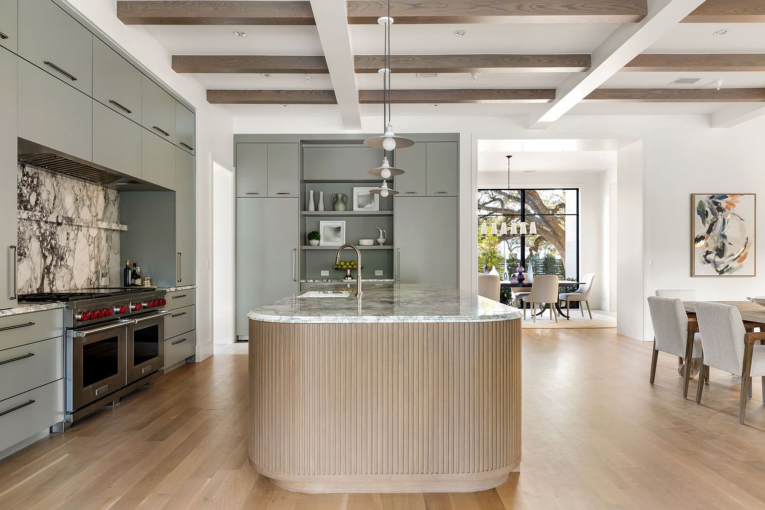 This is a bright and spacious kitchen featuring light gray cabinetry, a large island with a marble countertop and a wooden base, and stainless steel appliances. The kitchen also has a marble backsplash and wooden beams on the ceiling. The overall impression is modern and luxurious, with a focus on natural materials and light.