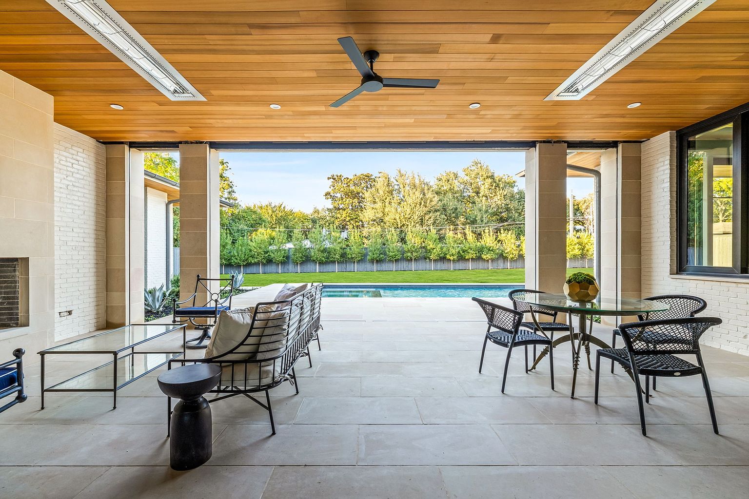 This image showcases a luxurious outdoor patio area, seamlessly blending indoor and outdoor living. The space features elegant patio furniture, including a seating area and a dining set, all set against a backdrop of a well-maintained lawn and a sparkling pool. The wooden ceiling with integrated lighting and a ceiling fan adds a touch of sophistication, while the open design provides unobstructed views of the backyard.
