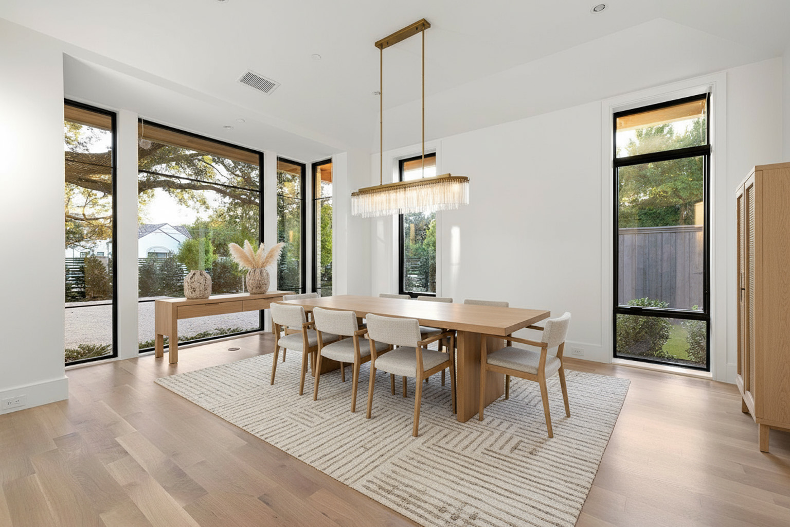 This is a bright and airy dining room with large windows offering natural light. The room features a long wooden dining table with upholstered chairs, a modern chandelier, and a neutral-toned area rug. The style is elegant and minimalist, perfect for a sophisticated dining experience.