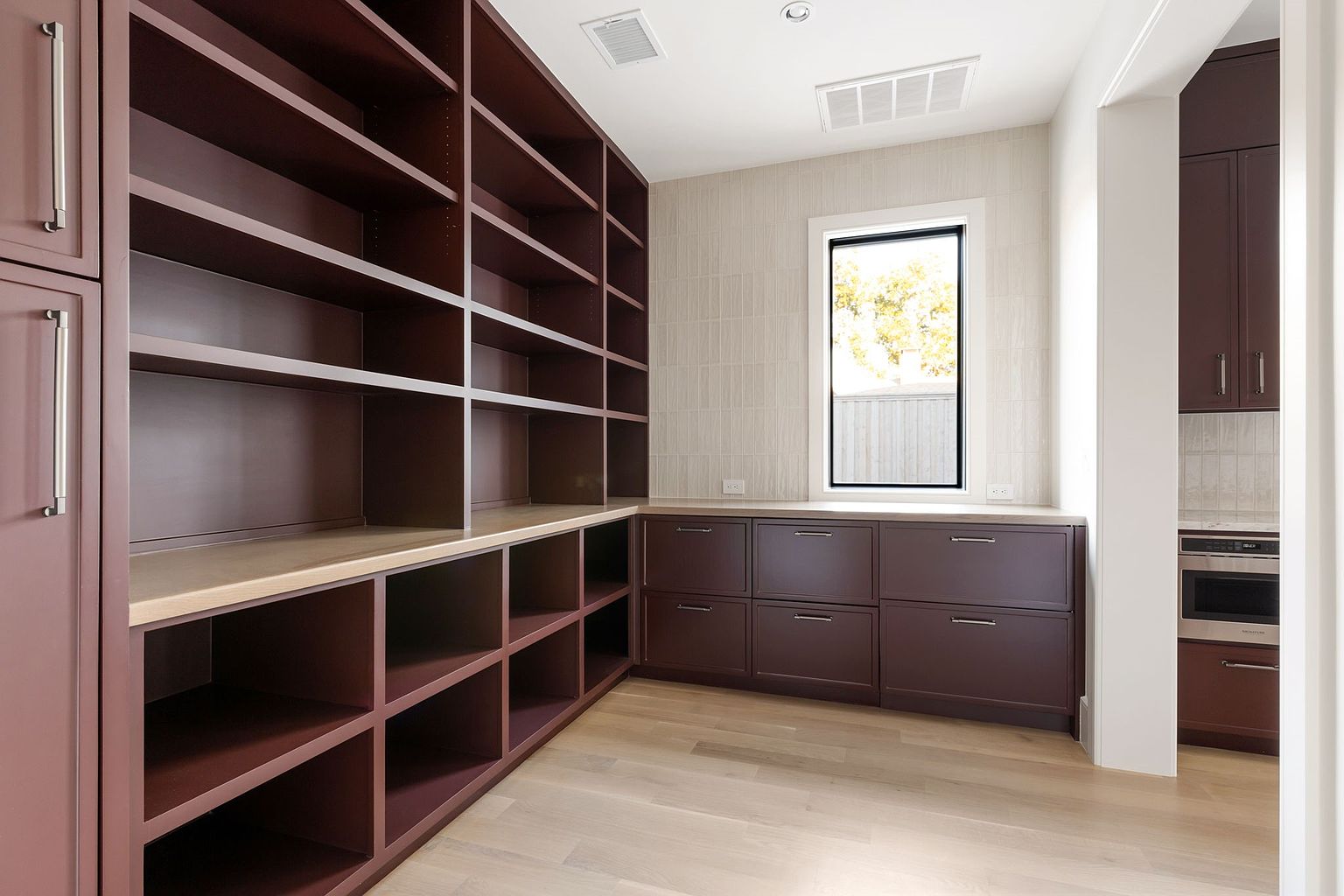 This is an interior shot of a well-organized pantry. The pantry features custom dark brown shelving and cabinets, providing ample storage space. A light-colored countertop runs along the base of the shelves, and a window provides natural light, enhancing the room's appeal.