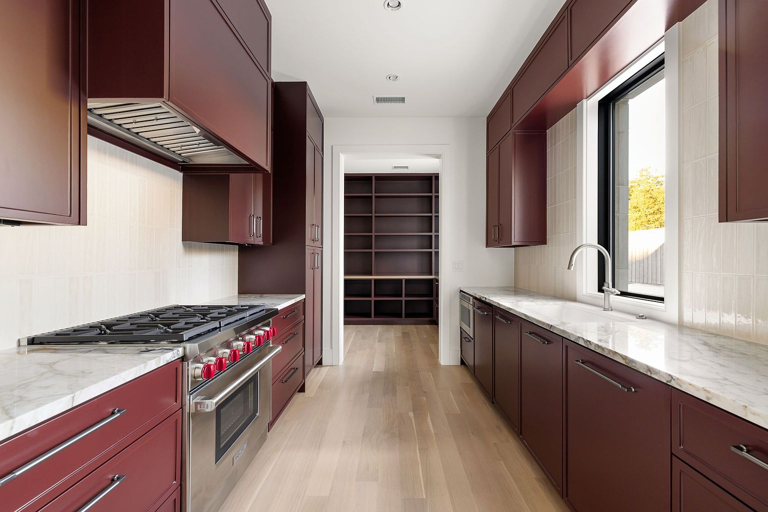 This is a galley kitchen with dark red cabinetry, marble countertops, and stainless steel appliances. The kitchen features a gas range with a stainless steel hood, a window over the sink, and a pantry at the end of the galley. The perspective is a straight-on shot, emphasizing the length of the kitchen and the storage space.