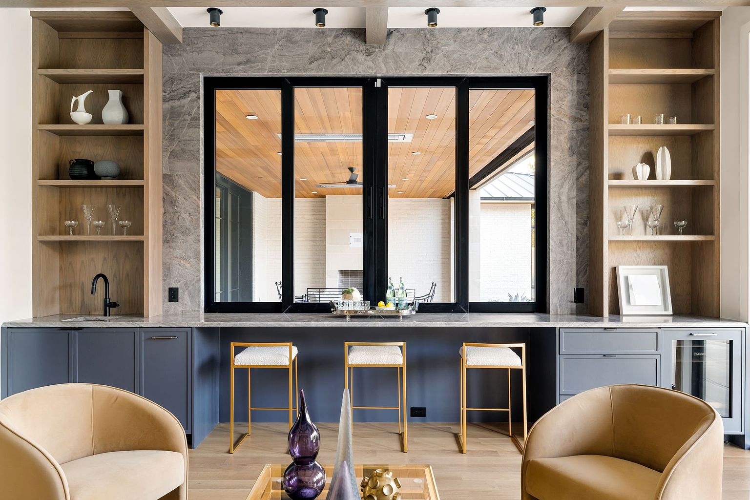 This interior shot showcases a stylish living room with a built-in bar area. The bar features dark blue cabinetry, marble backsplash, and open shelving, complemented by gold-framed bar stools. Large windows provide a view to an outdoor patio, and comfortable armchairs create an inviting seating arrangement.
