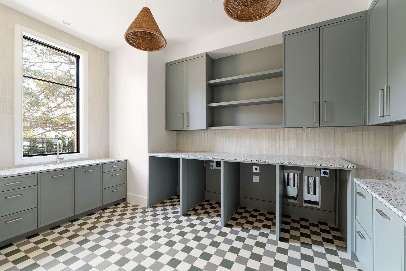 This is a well-organized laundry room featuring gray cabinetry with silver hardware and speckled countertops. The room includes open shelving, a window providing natural light, and a checkerboard tile floor. The space is functional and stylish, perfect for a modern home.
