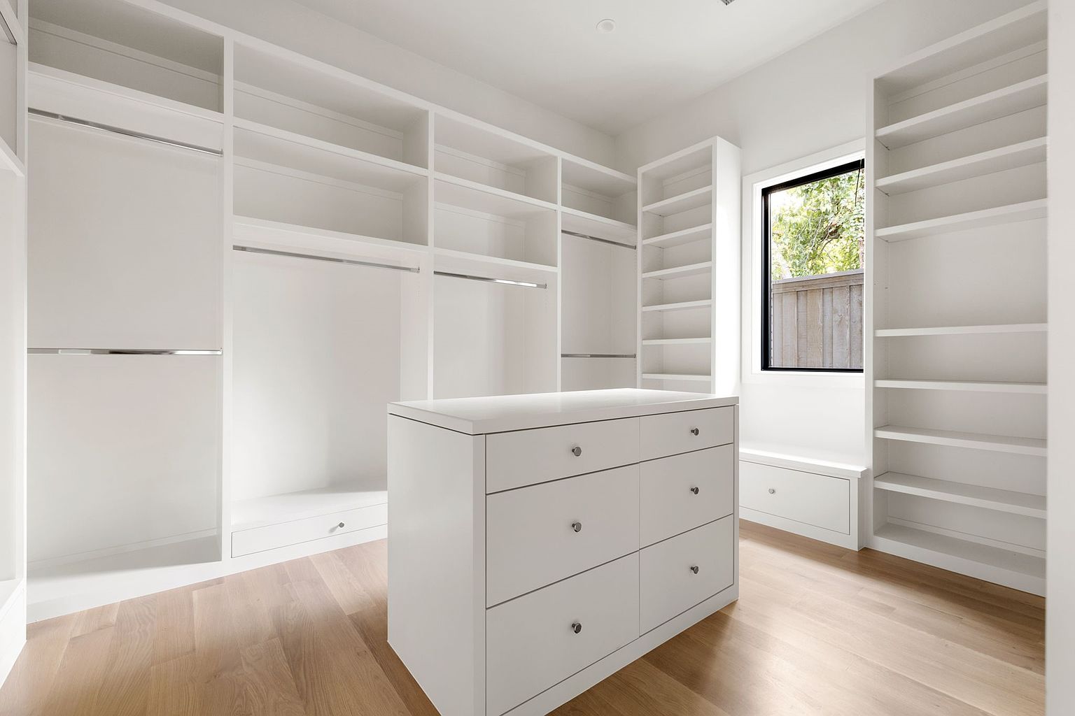 This is a bright and spacious walk-in closet featuring custom white shelving and cabinetry. A central island with drawers provides additional storage and countertop space. The light wood flooring and natural light from the window create a clean and luxurious feel, perfect for organizing and displaying clothing and accessories.
