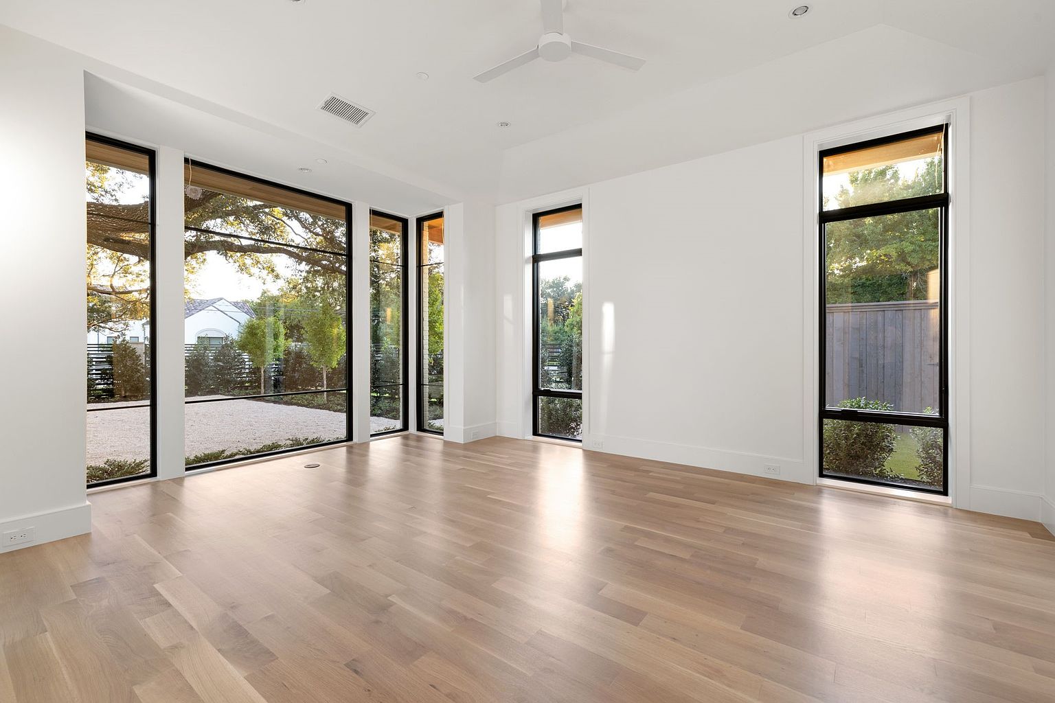 This is a spacious and bright living room with light hardwood floors and white walls. Large black-framed windows offer ample natural light and views of the exterior landscape. The room is modern and minimalist, creating a clean and airy atmosphere.