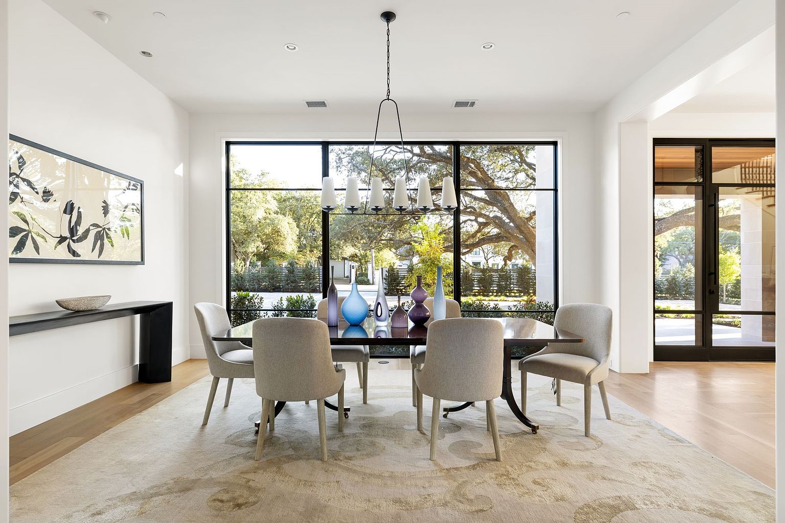 This is an interior shot of a modern dining room. The room features a large window with a view of the outdoors, a dining table with six chairs, and a modern chandelier. The color palette is neutral, with white walls and light-colored furniture, creating a bright and airy atmosphere.