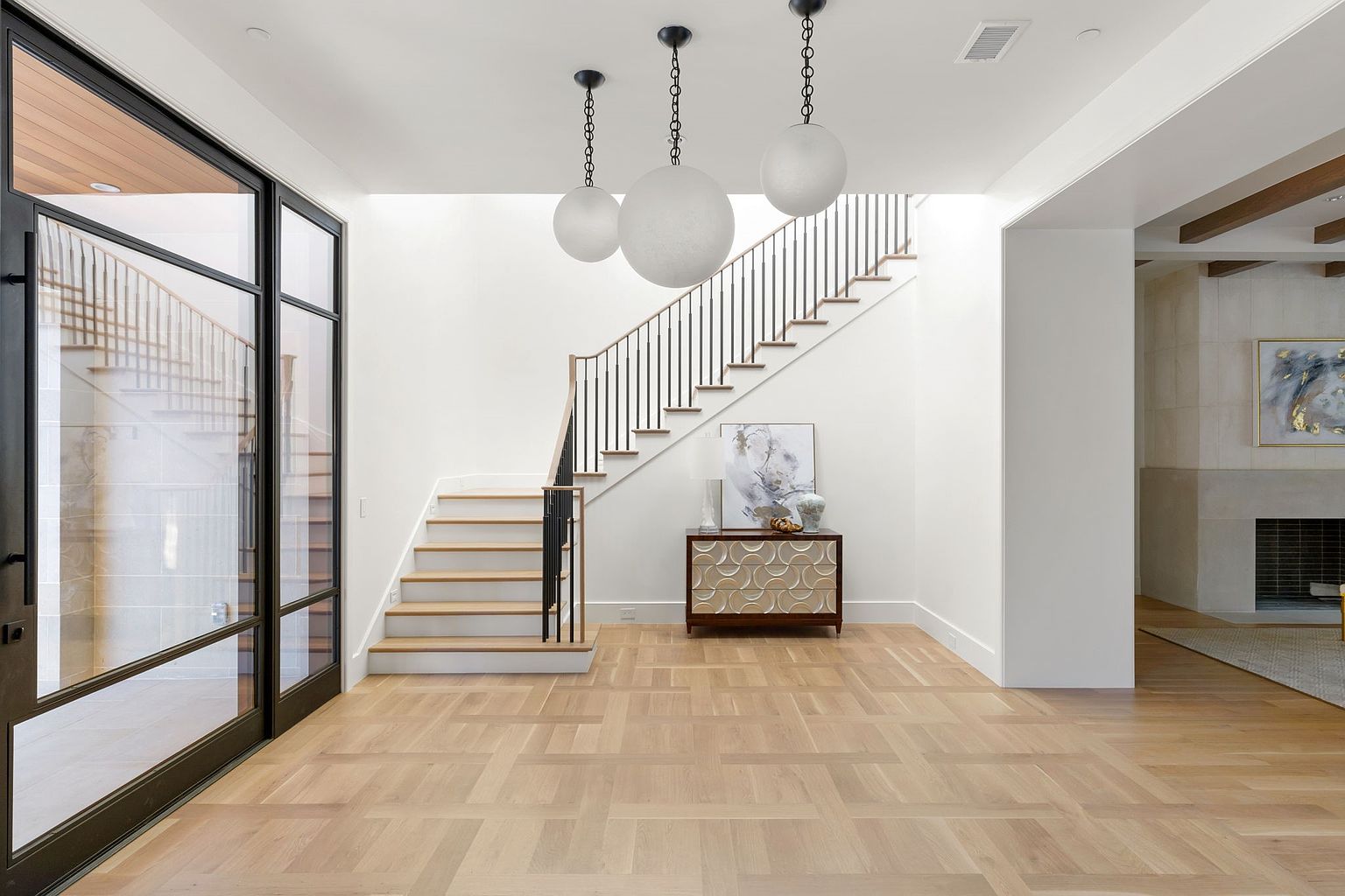This interior shot showcases a bright and modern entryway featuring a staircase with black iron railings and light wood steps. The flooring is a light-toned wood laid in a geometric pattern, and three spherical pendant lights hang from the ceiling, adding a touch of elegance. A console table with decorative details sits beneath a piece of artwork, enhancing the sophisticated ambiance of the space.