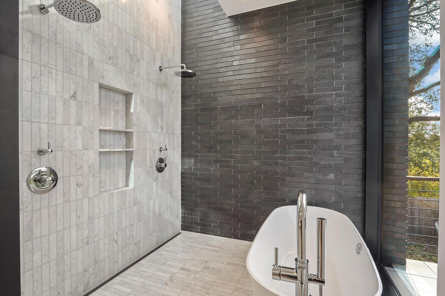 This is a modern primary bathroom featuring a walk-in shower with white marble tile and a built-in niche for storage. The adjacent wall is covered in dark gray brick-like tiles, contrasting with the white freestanding bathtub and its sleek chrome faucet. A large window provides natural light and a view of the outdoors, enhancing the spa-like atmosphere.