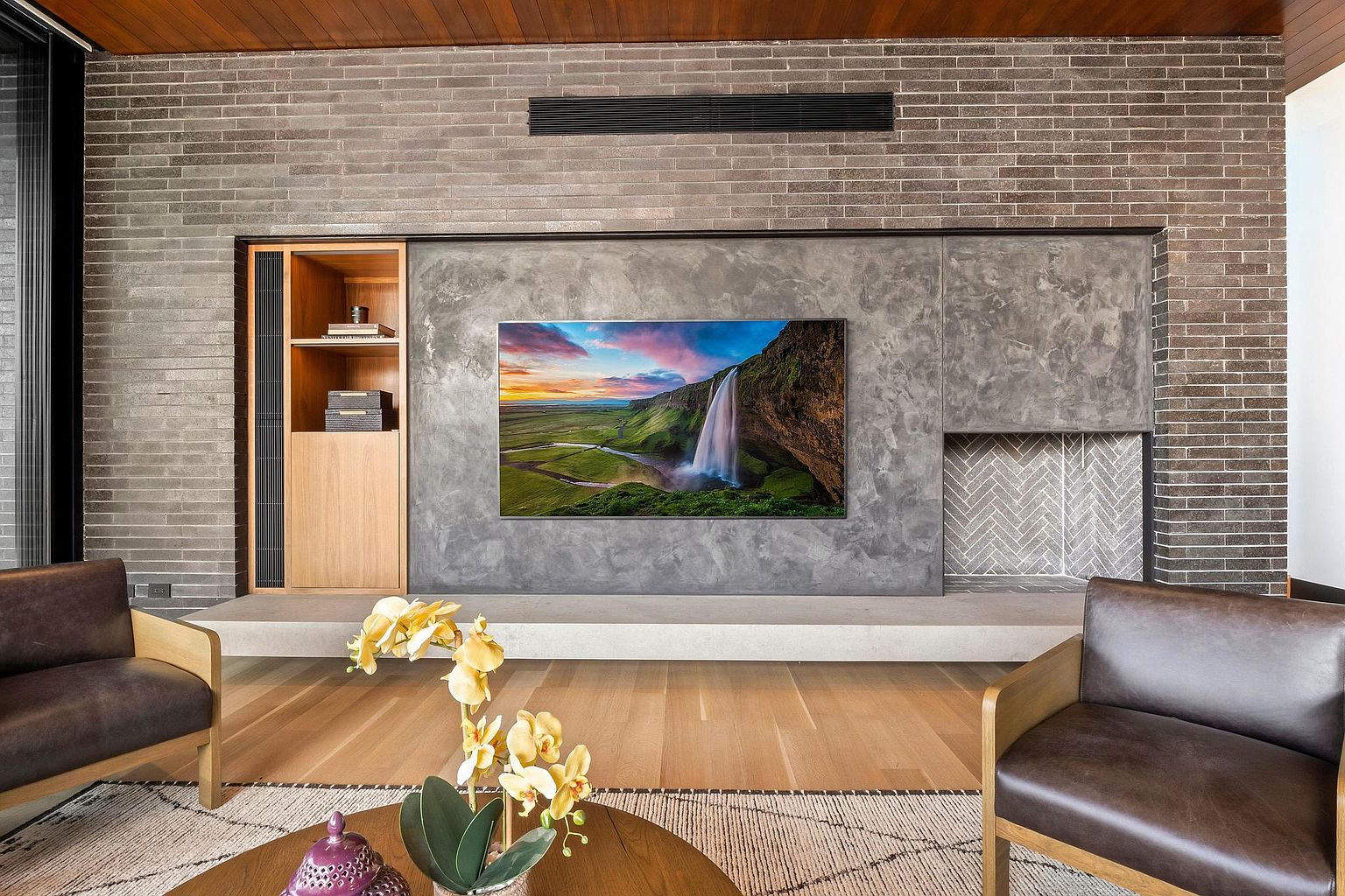 This living room showcases a modern aesthetic with a brick accent wall, a built-in entertainment center featuring a large television displaying a scenic landscape, and a decorative fireplace. Two leather armchairs flank a round coffee table with floral decor, set upon a patterned area rug and hardwood flooring. The room exudes a sophisticated and comfortable ambiance.