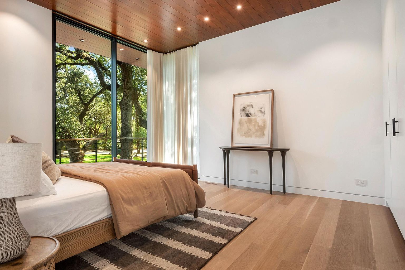 This is a bright and airy primary bedroom featuring a large window with a view of lush greenery, a wooden ceiling with recessed lighting, and hardwood floors. The room is minimally furnished with a bed, a console table with artwork, and a striped rug, creating a serene and modern atmosphere. The perspective is from the corner of the room, showcasing the bed and the view.