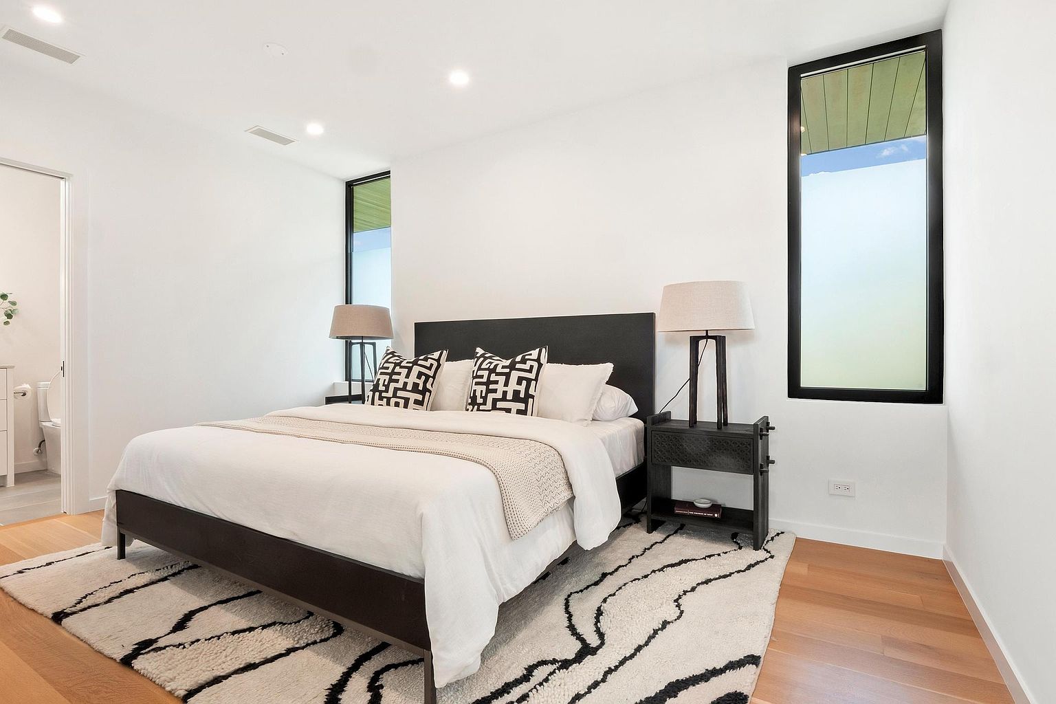 This is a primary bedroom featuring a dark-framed bed with white linens and patterned pillows, flanked by matching dark wood nightstands and lamps. The room has white walls, hardwood floors, and a black-framed window that provides natural light. A patterned rug adds texture to the space, creating a modern and inviting atmosphere.