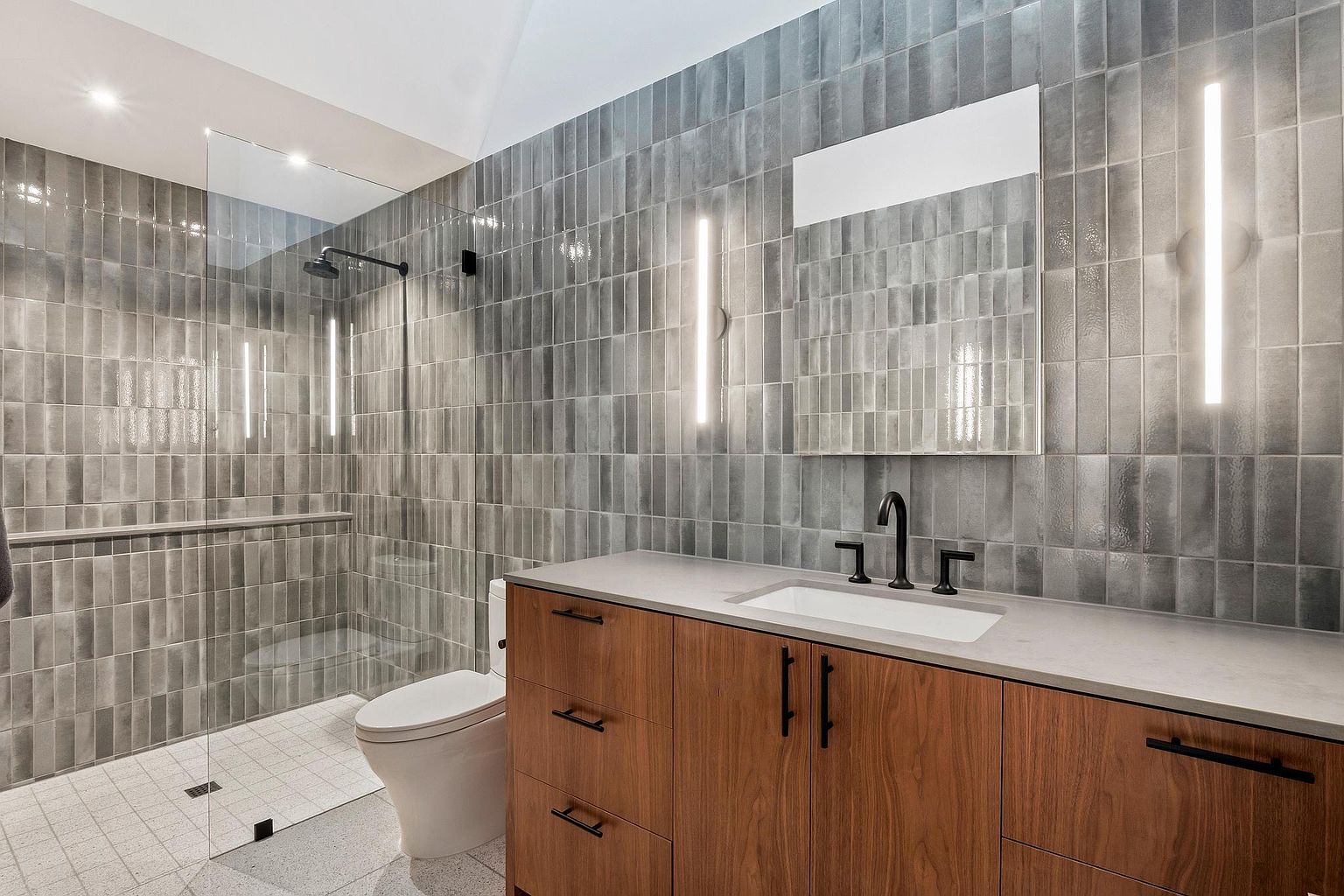 This is a modern primary bathroom featuring gray vertical tiles on the walls and a light-colored, speckled floor. A glass-enclosed shower is on the left, and a wooden vanity with a light countertop and black hardware is on the right. The bathroom is well-lit with vertical light fixtures flanking a mirror above the vanity, creating a clean and contemporary aesthetic.