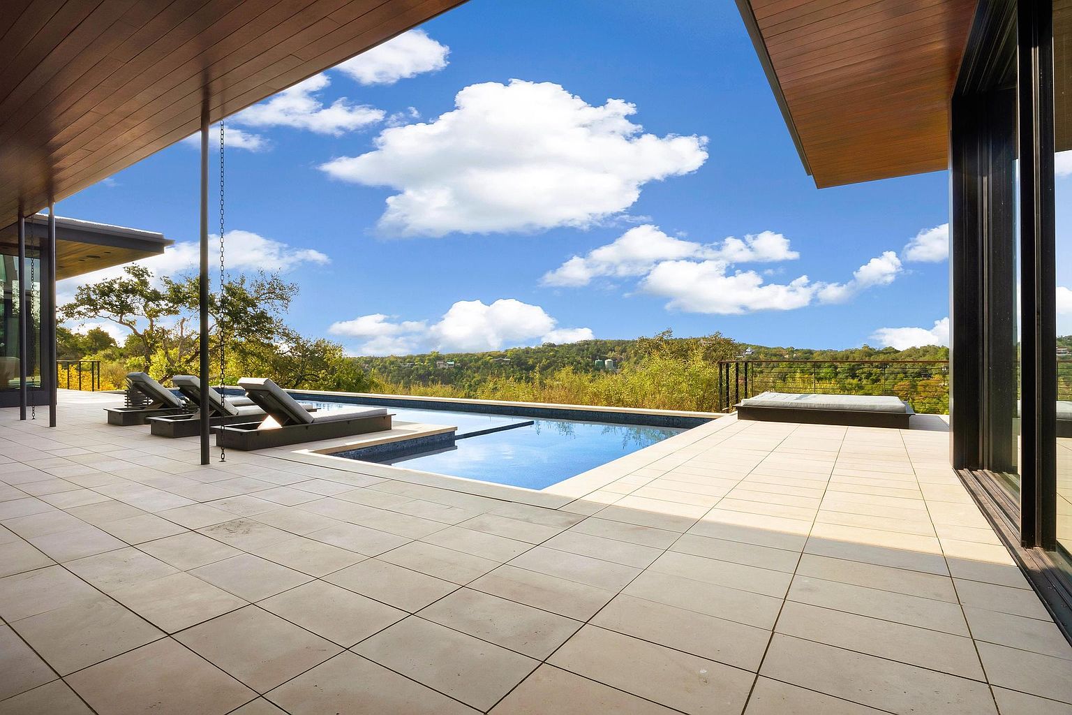 This image showcases a luxurious outdoor pool area with a modern design. The pool is surrounded by a spacious tiled patio, lounge chairs, and lush greenery in the background. The open-air structure provides a stunning view of the landscape and sky, creating a serene and inviting atmosphere.