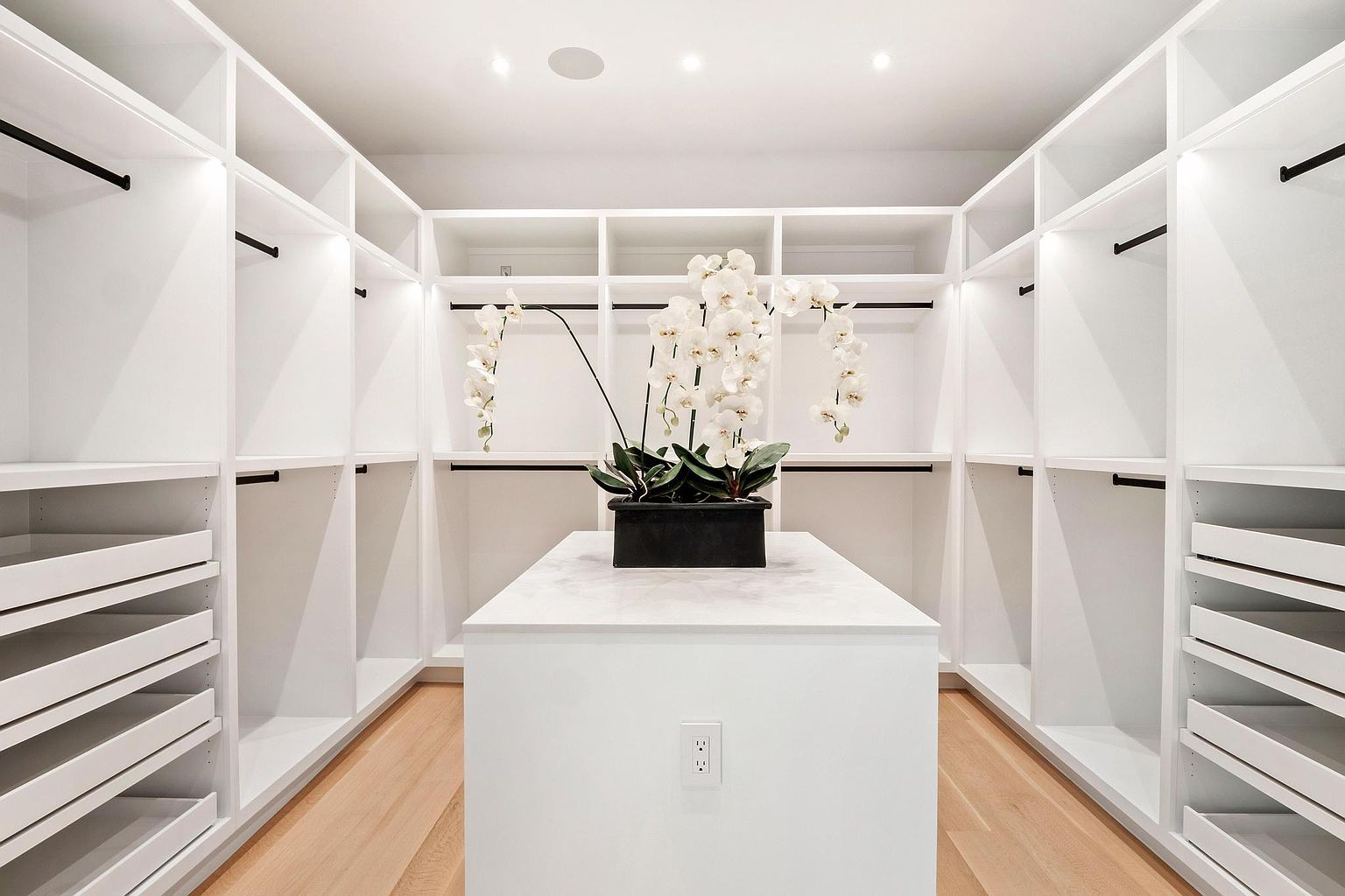 This is a well-organized walk-in closet featuring custom white shelving and drawers. A central island with a light-colored countertop provides additional storage and display space, topped with a floral arrangement. The light hardwood flooring and recessed lighting create a bright and luxurious atmosphere.