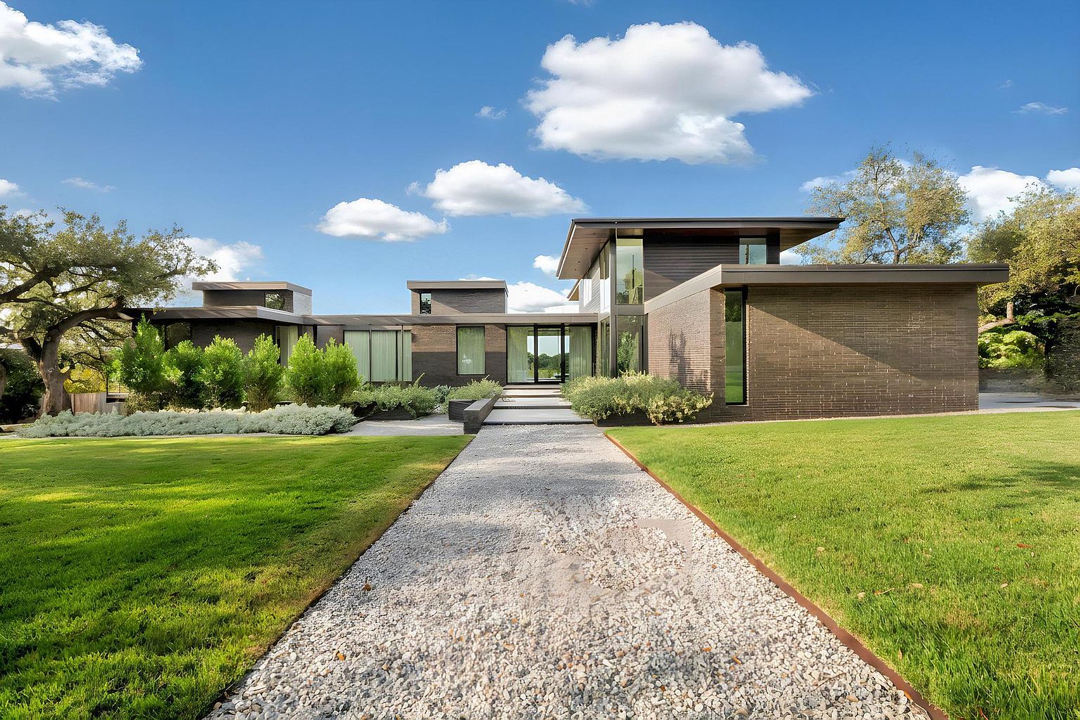 This image showcases the front exterior of a modern, single-story home with a striking architectural design. The house features a dark brick facade, large windows, and a flat roof. A gravel pathway leads to the entrance, flanked by well-manicured lawns and landscaping, creating an inviting and sophisticated curb appeal.