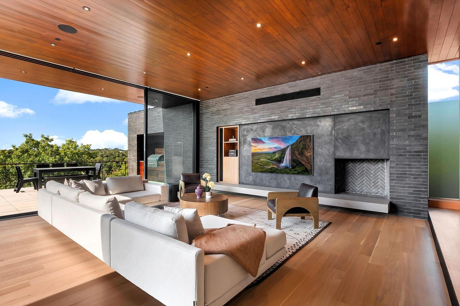 This is a modern living room featuring a large white sectional sofa, a wooden ceiling, and hardwood floors. A large sliding glass door opens to an outdoor patio. The focal point is a gray brick wall with a built-in entertainment center and a fireplace, creating a sophisticated and inviting atmosphere.