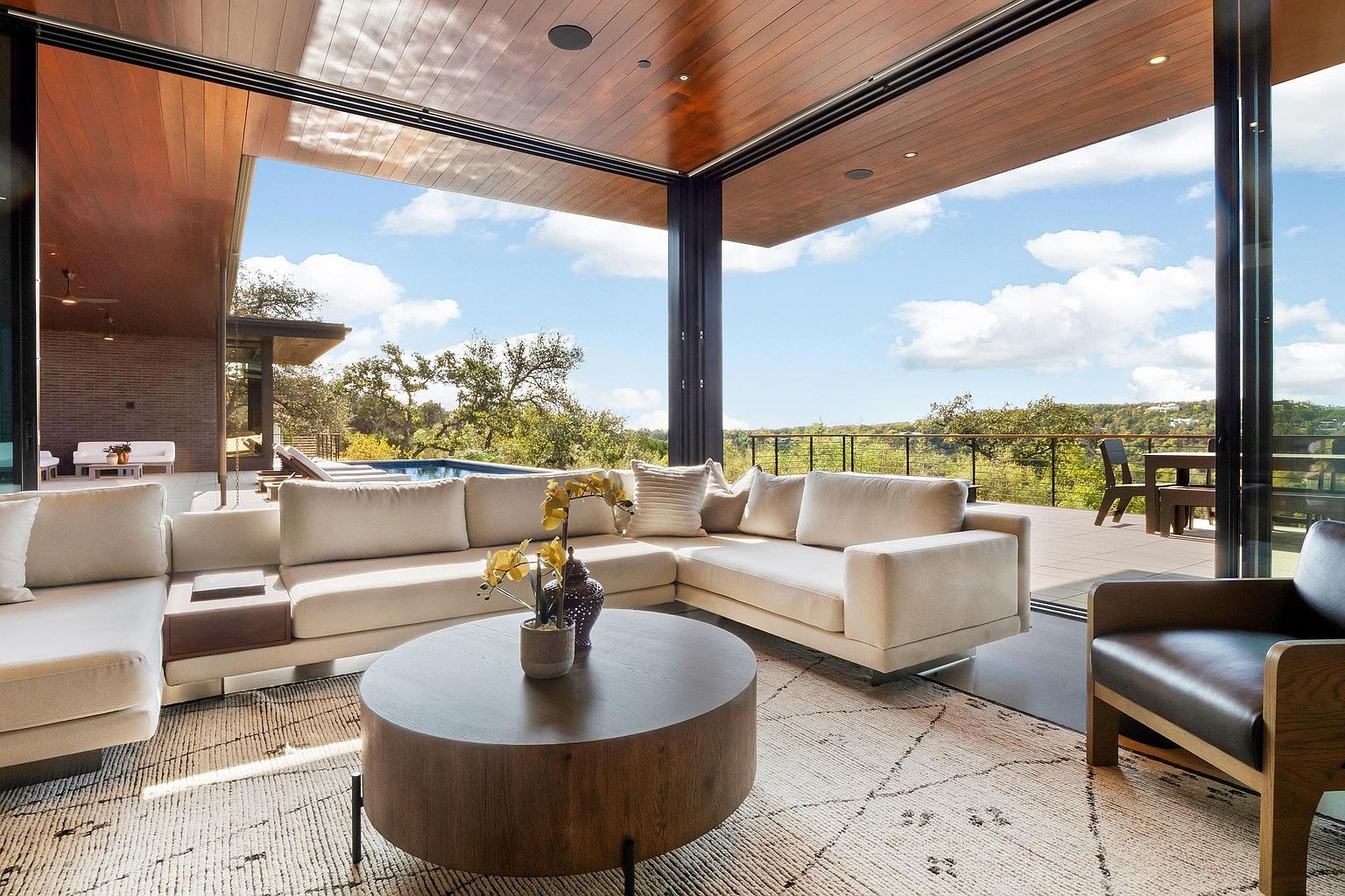 This is a bright and airy living room featuring a large sectional sofa, a round wooden coffee table, and a neutral-toned rug. The room opens to an outdoor deck with a view of the surrounding landscape through large sliding glass doors, creating a seamless indoor-outdoor living experience. The wood ceiling adds warmth to the modern design.