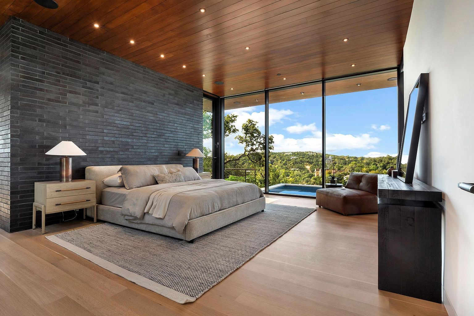 This is a luxurious primary bedroom featuring a modern design with a dark brick accent wall and floor-to-ceiling windows offering stunning views of the landscape. The room is furnished with a large bed, nightstands with lamps, a console table with a mirror, and a comfortable armchair, all contributing to a serene and upscale atmosphere. The hardwood floors and recessed lighting add to the room's elegance and appeal.