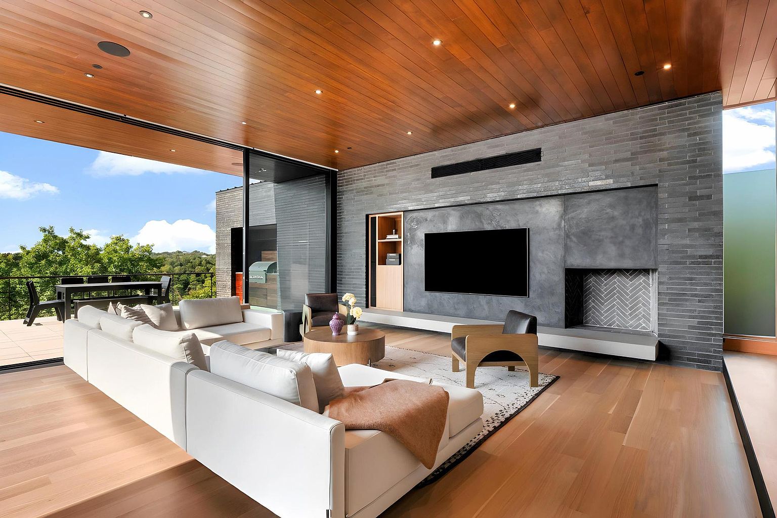 This is a modern living room featuring a large white sectional sofa, a round coffee table, and a sleek entertainment center with a flat-screen TV and a fireplace. The room has hardwood floors, a wood-paneled ceiling, and a large sliding glass door that opens to an outdoor balcony. The overall style is contemporary and luxurious, with a focus on clean lines and natural materials.