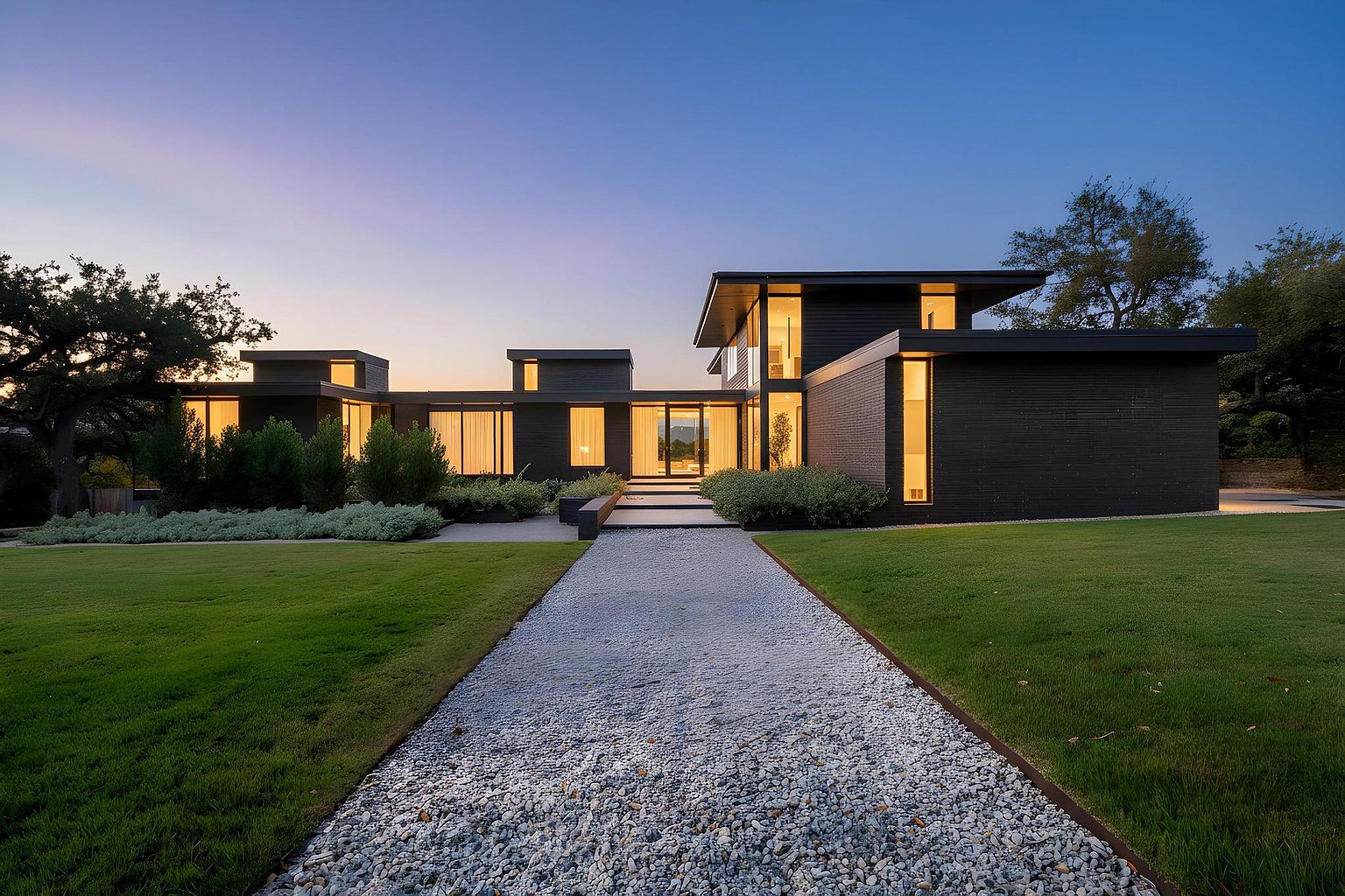 This image showcases the front exterior of a modern, single-story home at dusk. A gravel pathway leads to the entrance, flanked by manicured lawns and garden beds. The house features dark siding, large windows that glow with interior light, and a flat roof design, creating a sleek and sophisticated aesthetic.