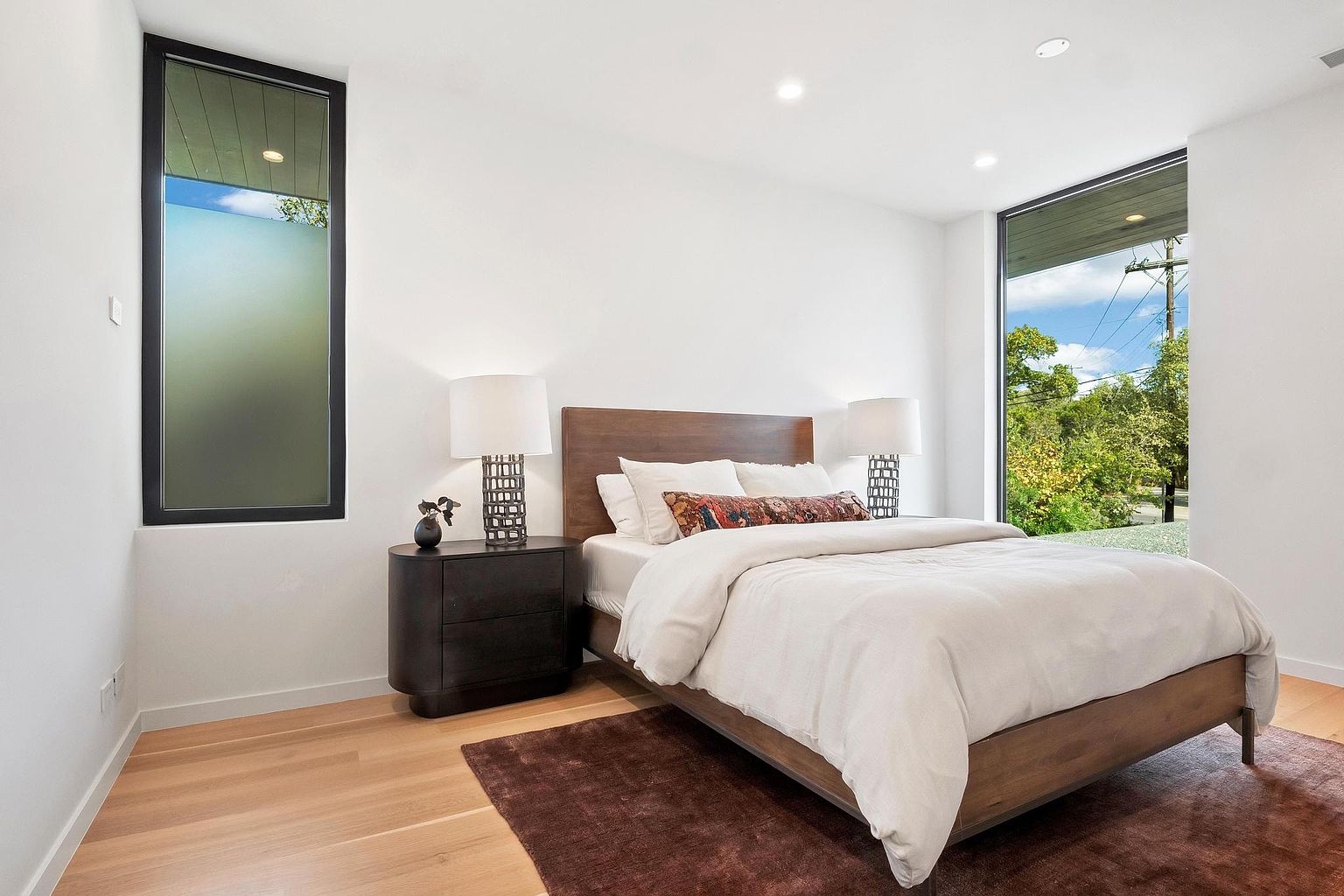 This is a bright and airy primary bedroom featuring a wooden bed frame with a white duvet and decorative pillows. The room has white walls, light wood flooring, and two large windows that provide natural light and views of the outdoors. A dark wood nightstand with lamps on either side of the bed adds a touch of elegance.