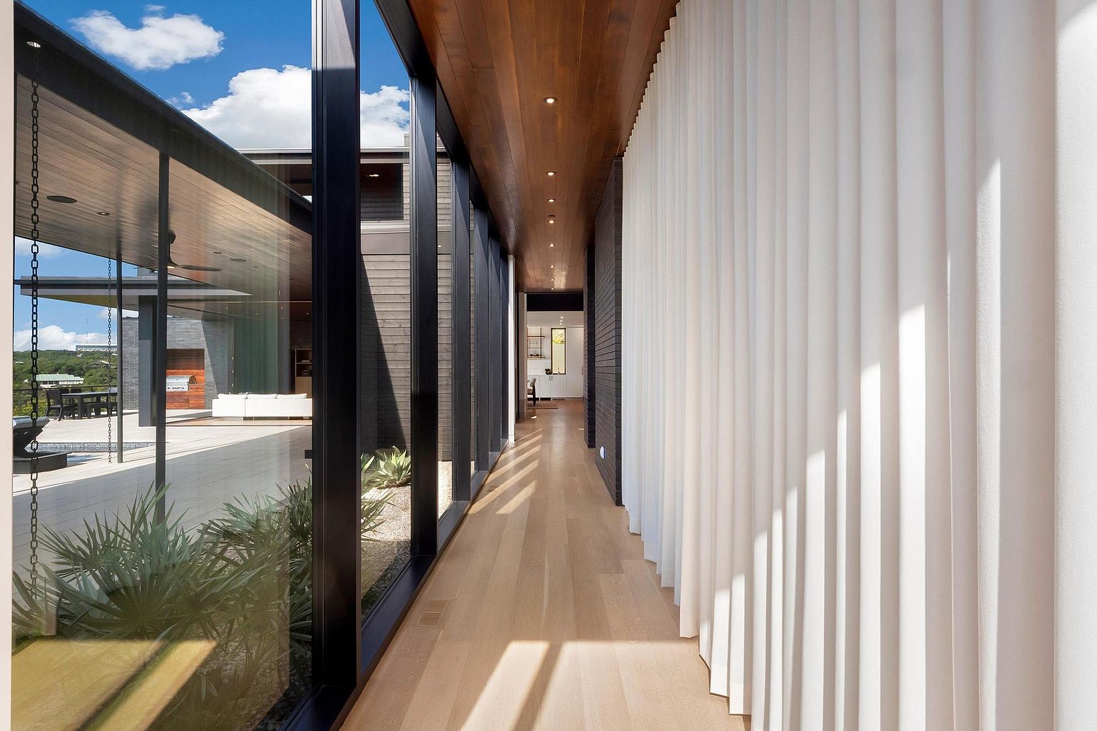 This interior shot showcases a modern hallway with a striking combination of natural light and sleek design. A wall of floor-to-ceiling windows on one side offers views of an outdoor patio and greenery, while the opposite wall features textured white panels. The hallway is illuminated by recessed lighting in the wooden ceiling, and the light wood flooring adds warmth to the space, creating a bright and inviting atmosphere.