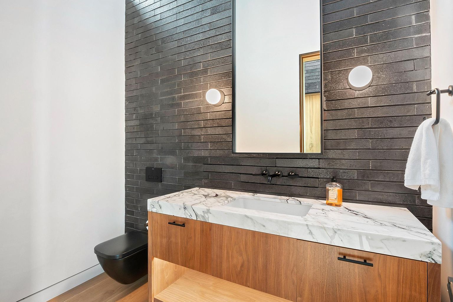 This is a modern guest bathroom featuring a sleek black toilet and a vanity with a marble countertop and wood cabinetry. The wall is covered in dark, horizontally laid brick tiles, complemented by minimalist lighting fixtures and black hardware. The overall impression is stylish and contemporary.