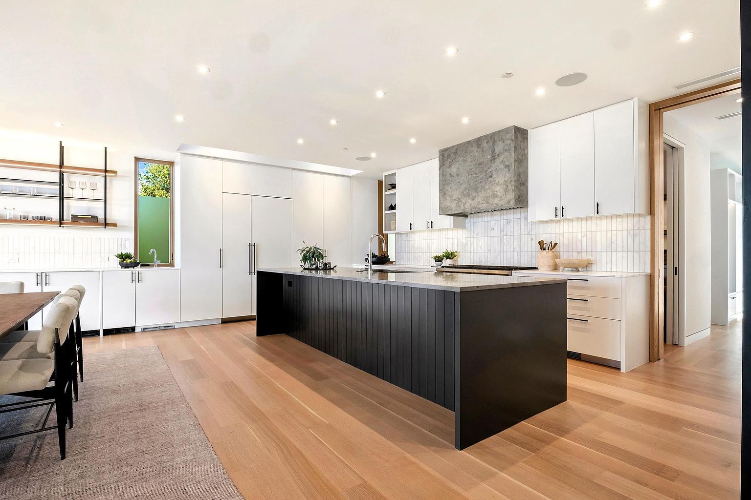 This is a bright and modern kitchen featuring a large dark-colored island with a light countertop, white cabinetry, and stainless steel appliances. The kitchen has a clean, minimalist design with recessed lighting and a light wood floor. The perspective is from a wide angle, capturing the island and the surrounding cabinetry.