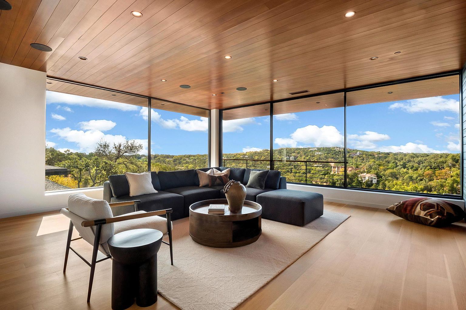 This is a bright and modern living room featuring floor-to-ceiling windows that offer panoramic views of the surrounding landscape. The room is furnished with a comfortable sectional sofa, a round coffee table, and a stylish accent chair, all arranged on a neutral-toned area rug. The wood-paneled ceiling adds warmth and character to the space, creating an inviting and luxurious atmosphere.