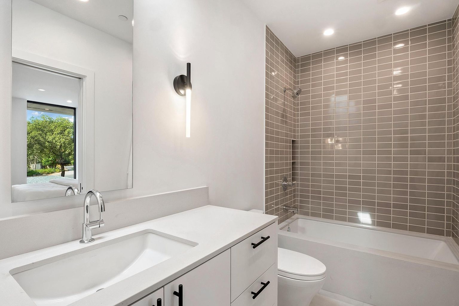 This is a well-lit bathroom featuring a white vanity with black hardware, a modern faucet, and a large mirror reflecting an outdoor scene. The bathroom includes a toilet and a bathtub with gray tiled walls, creating a clean and contemporary aesthetic. The perspective is from the doorway, showcasing the entire bathroom layout.