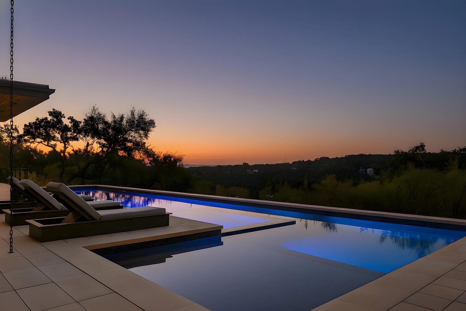 This image showcases a luxurious infinity pool at dusk, featuring built-in lighting that casts a serene blue glow. Lounge chairs are positioned poolside, offering a relaxing space to enjoy the panoramic view of the surrounding landscape and the colorful sunset. The pool's design seamlessly blends with the natural environment, creating a tranquil and upscale ambiance.