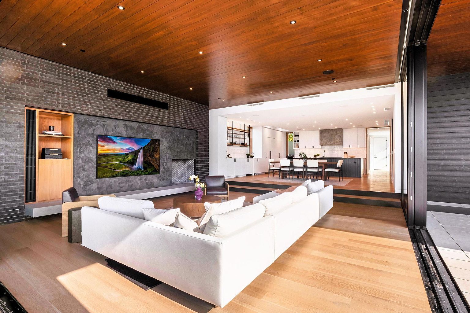 This is a spacious and modern living room that seamlessly blends indoor and outdoor living. The room features a large white sectional sofa, a wooden ceiling with recessed lighting, and a brick accent wall with a built-in entertainment center. A large sliding glass door opens to an outdoor area, creating an open and airy atmosphere.