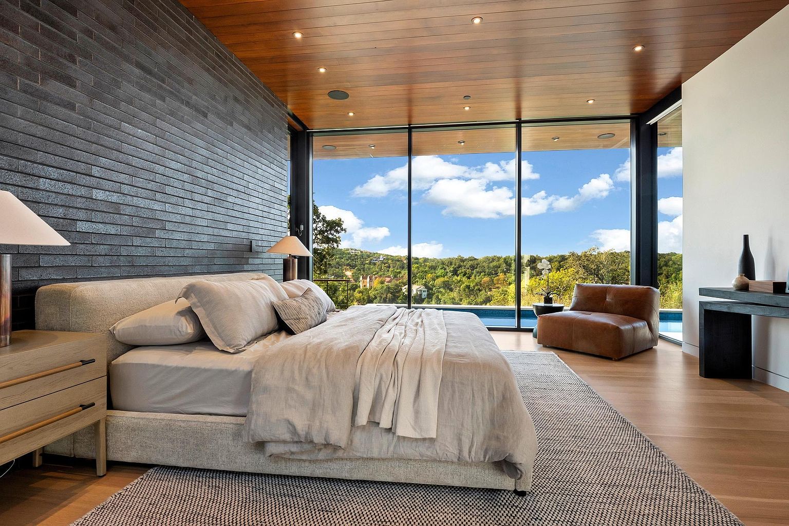 This is a luxurious primary bedroom featuring a large bed with neutral linens, a textured accent wall, and floor-to-ceiling windows offering a stunning view of the surrounding landscape and pool. The room is well-lit with natural light and features hardwood floors and a comfortable lounge chair, creating a serene and inviting atmosphere.