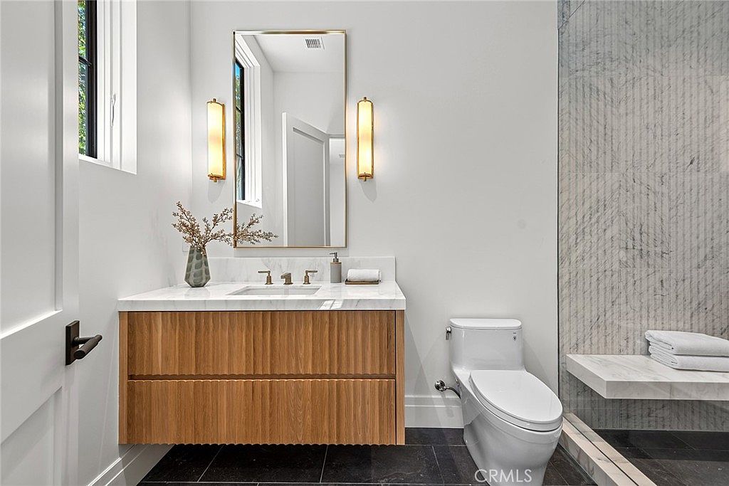 This modern bathroom features a floating vanity with a textured wood finish, topped with a clean white marble countertop and a sleek undermount sink. A tall, rectangular mirror is flanked by two elegant vertical wall sconces, while a walk-in shower with marble-tiled walls and a built-in bench sits to the right. The space is finished with dark floor tiles and crisp white walls, creating a sophisticated and minimalist aesthetic.