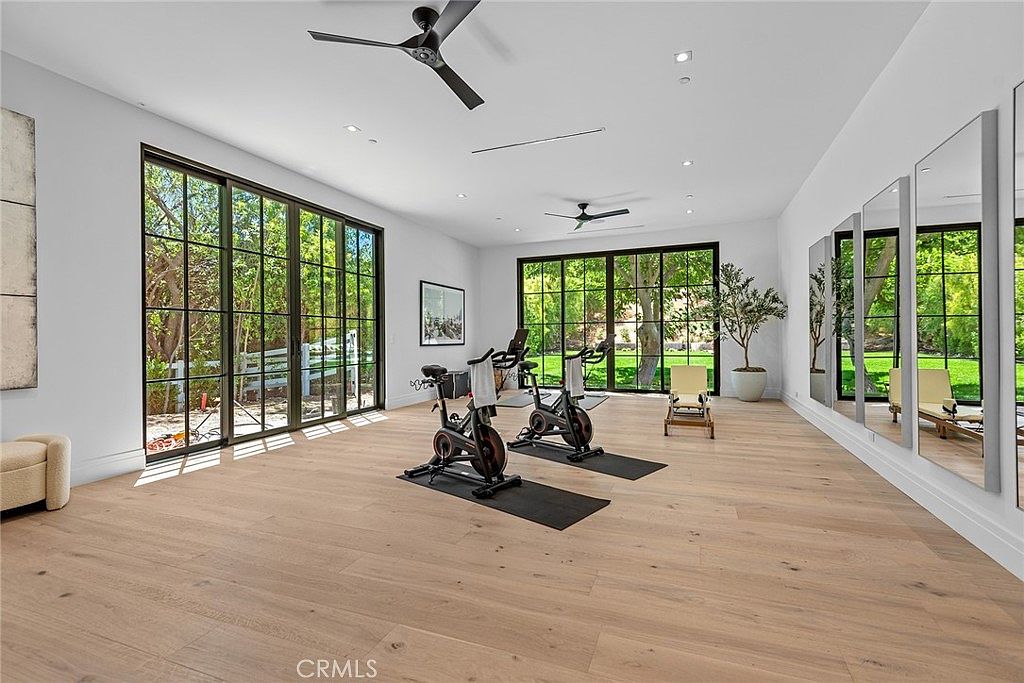 This spacious and bright home gym features light-toned hardwood flooring, high ceilings with recessed lighting, and two ceiling fans. The room is equipped with two stationary exercise bikes on individual mats and a wall of large mirrors, while expansive floor-to-ceiling glass doors provide abundant natural light and views of the lush greenery outside. The minimalist design creates an airy, professional, and inviting atmosphere for a workout.