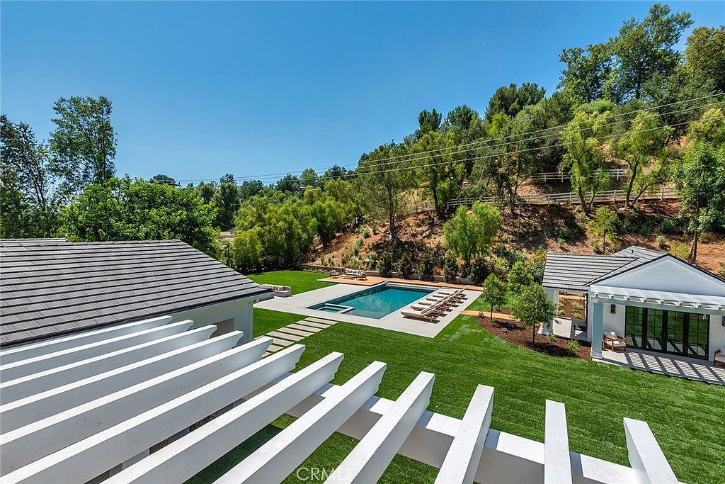 This elevated perspective captures a beautifully landscaped backyard featuring a pristine rectangular swimming pool as the central focal point. The scene includes a lush green lawn, a modern pergola structure in the foreground, and a charming pool house, all set against a backdrop of mature trees and a hillside. The overall impression is one of luxury, tranquility, and high-end outdoor living.