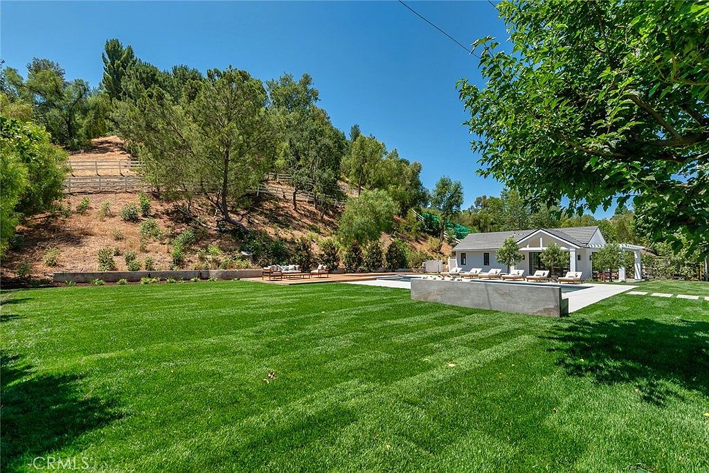 This expansive backyard features a meticulously manicured green lawn that leads to a modern pool area and a detached pool house. The property is set against a scenic, sloped hillside with mature trees and a white fence, creating a private and serene atmosphere. The scene is captured from a low-angle perspective, emphasizing the vastness of the outdoor space and the seamless integration of luxury landscaping with the natural environment.