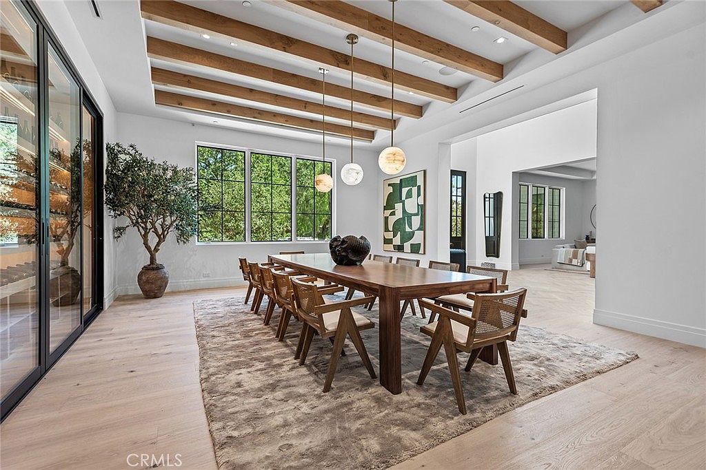 This elegant dining room features a long wooden table surrounded by modern woven chairs, set atop a large, textured area rug. The space is defined by a striking coffered ceiling with exposed wooden beams and three contemporary globe pendant lights. To the left, a glass-enclosed wine cellar adds a sophisticated touch, while large windows provide ample natural light and a view of the lush greenery outside.